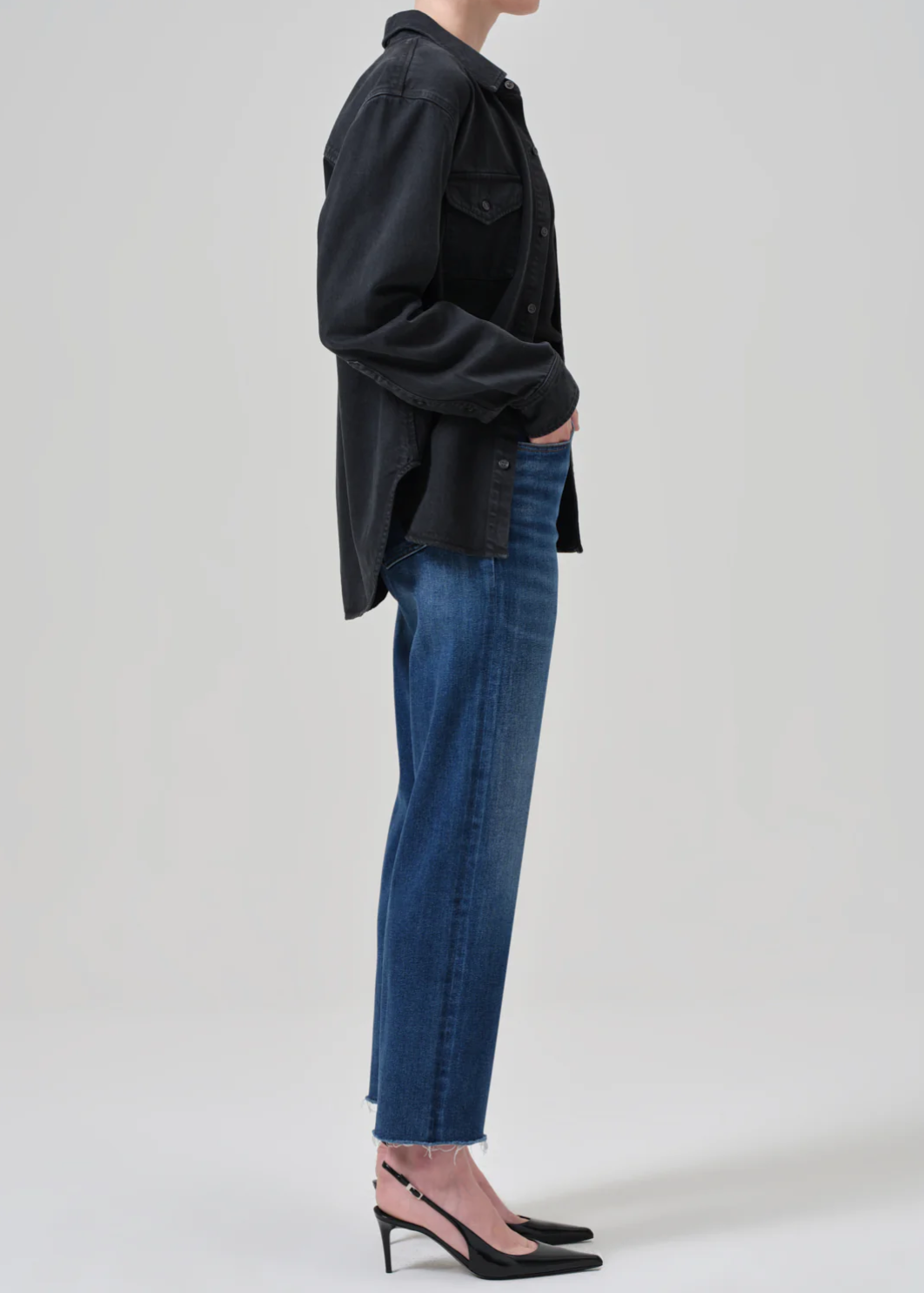 A person is standing against a plain background, facing to the left. They are wearing a loose-fitting, black button-up shirt, light blue Palma Straight jeans in Ambry by Citizens Of Humanity/AGOLDE with frayed hems, and black pointed-toe heels with ankle straps. Their hands are in their pockets.