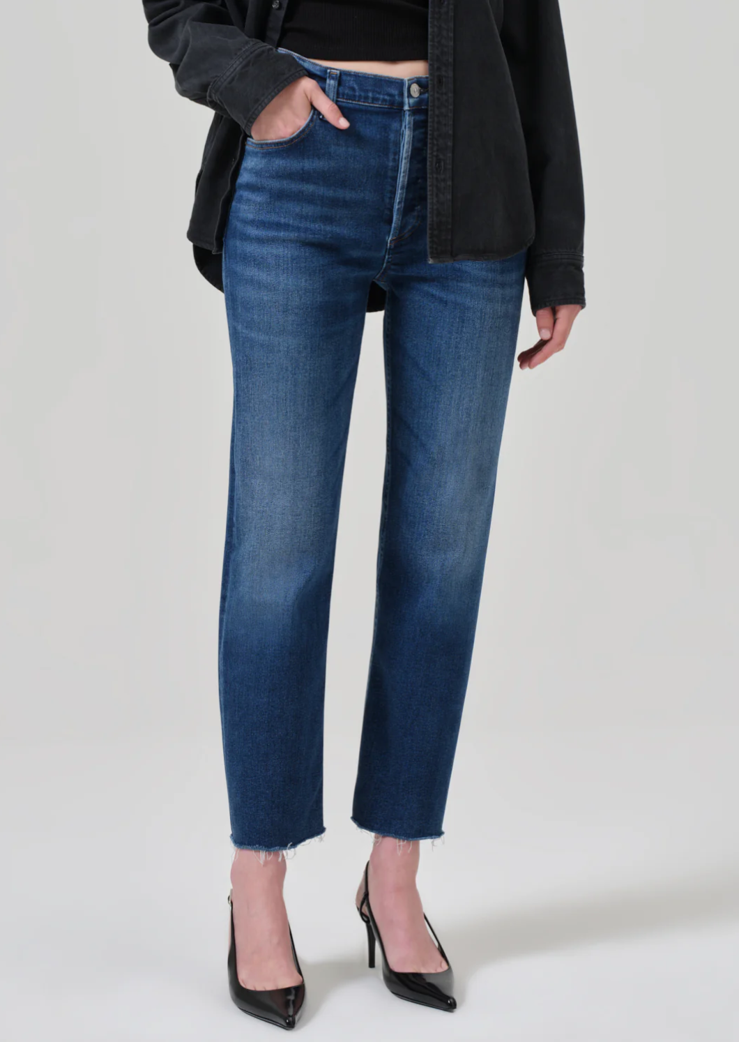 A person stands against a plain background, showcasing a casual outfit featuring the Palma Straight jeans in Ambry from Citizens Of Humanity/AGOLDE with frayed hems, a black top, and a dark jacket. They also wear black pointed-toe heels. Their left hand is in their pocket.