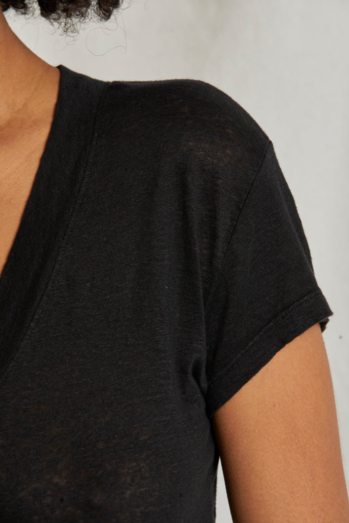 Close-up of a person wearing the Rosie V-neck by Perfectwhitetee. The image highlights part of the person's shoulder, upper arm, and neck, showcasing the texture and fit of this classic black T-shirt. The background is plain and light-colored.