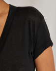 Close-up of a person wearing the Rosie V-neck by Perfectwhitetee. The image highlights part of the person's shoulder, upper arm, and neck, showcasing the texture and fit of this classic black T-shirt. The background is plain and light-colored.