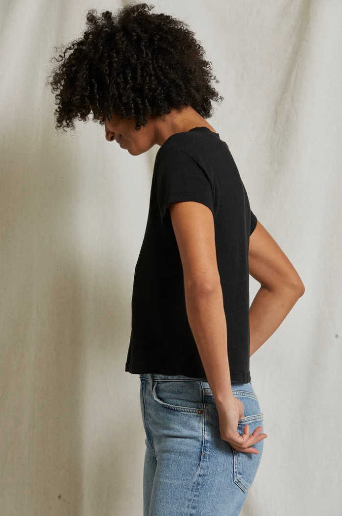 A person with curly hair, wearing a Rosie V-neck by Perfectwhitetee and blue jeans, stands in profile against a neutral background. Their head is slightly bowed, and they have one hand resting behind their back.