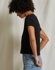 A person with curly hair, wearing a Rosie V-neck by Perfectwhitetee and blue jeans, stands in profile against a neutral background. Their head is slightly bowed, and they have one hand resting behind their back.