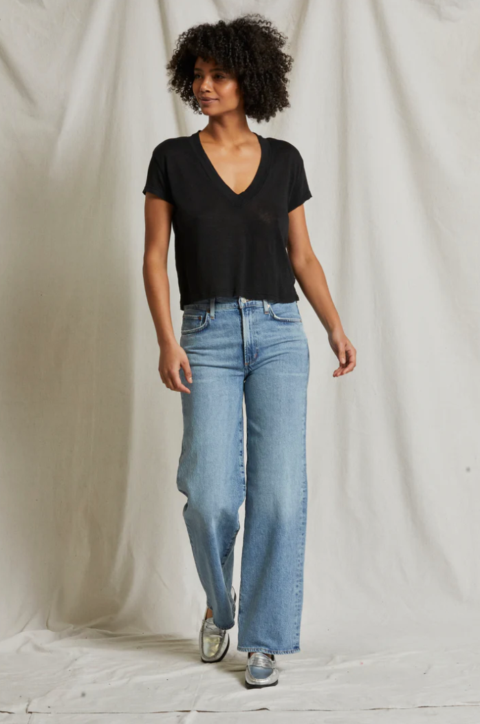 A person with curly hair stands against a light backdrop. They are wearing a Perfectwhitetee Rosie V-neck short sleeve top and light blue, high-waisted, wide-leg jeans. They also have on silver sneakers. The individual is looking off to the side with a relaxed expression.