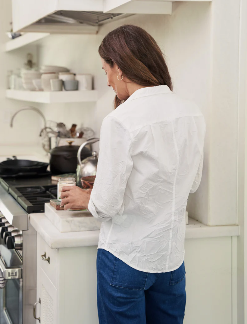 A woman with long brown hair, dressed in a 100% cotton white BARRY CRINKLE shirt from Frank & Eileen and blue jeans, stands in a kitchen with her back to the camera. She is preparing something on the counter near a stove with pots and a kettle in the background.