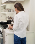 A woman with long brown hair, dressed in a 100% cotton white BARRY CRINKLE shirt from Frank & Eileen and blue jeans, stands in a kitchen with her back to the camera. She is preparing something on the counter near a stove with pots and a kettle in the background.