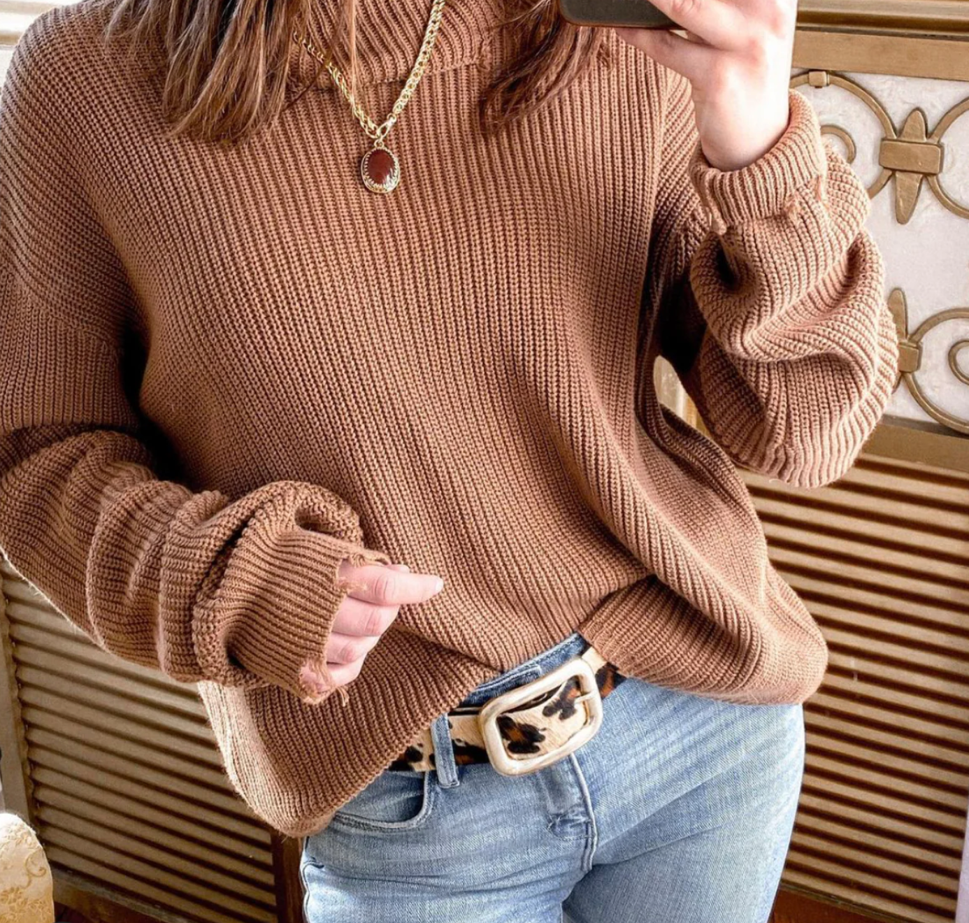 A person is wearing a brown knit sweater, blue jeans, and the Julianne Leopard Belt by Streets Ahead Inc, which features a brown and white patterned buckle. The person is taking a selfie with the phone partially visible. They are also wearing a necklace, and the background includes patterned wooden accents.