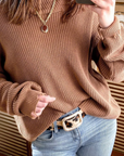 A person is wearing a brown knit sweater, blue jeans, and the Julianne Leopard Belt by Streets Ahead Inc, which features a brown and white patterned buckle. The person is taking a selfie with the phone partially visible. They are also wearing a necklace, and the background includes patterned wooden accents.