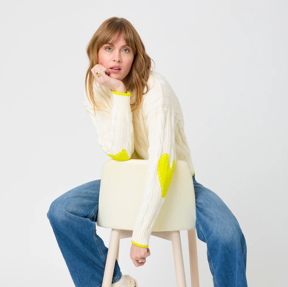 A person with blonde hair, donning the Kerri Rosenthal Maxwork Cable Sweater—a cozy white sweater adorned with yellow heart elbow patches—is seated on a white stool. They are wearing blue jeans and lean forward with their chin resting on their hand, looking directly at the camera against a plain white background.