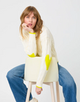 A person with blonde hair, donning the Kerri Rosenthal Maxwork Cable Sweater—a cozy white sweater adorned with yellow heart elbow patches—is seated on a white stool. They are wearing blue jeans and lean forward with their chin resting on their hand, looking directly at the camera against a plain white background.