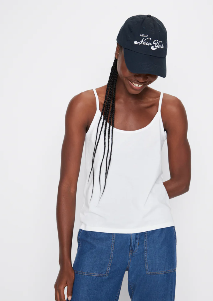 A person smiles with their head tilted down, wearing *The New York Kap* by Kule, a white tank top, and blue jeans. They have long braided hair and are standing against a plain white background, proudly embodying the spirit of a true New Yorker.