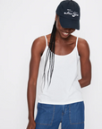 A person smiles with their head tilted down, wearing *The New York Kap* by Kule, a white tank top, and blue jeans. They have long braided hair and are standing against a plain white background, proudly embodying the spirit of a true New Yorker.