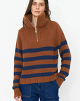 A person with long hair wears The Matey, a chunky cotton knit sweater by Kule, featuring brown and navy blue stripes with a high collar and a quarter zip. Paired with blue jeans, they stand against a plain white background, one hand casually in their pocket.