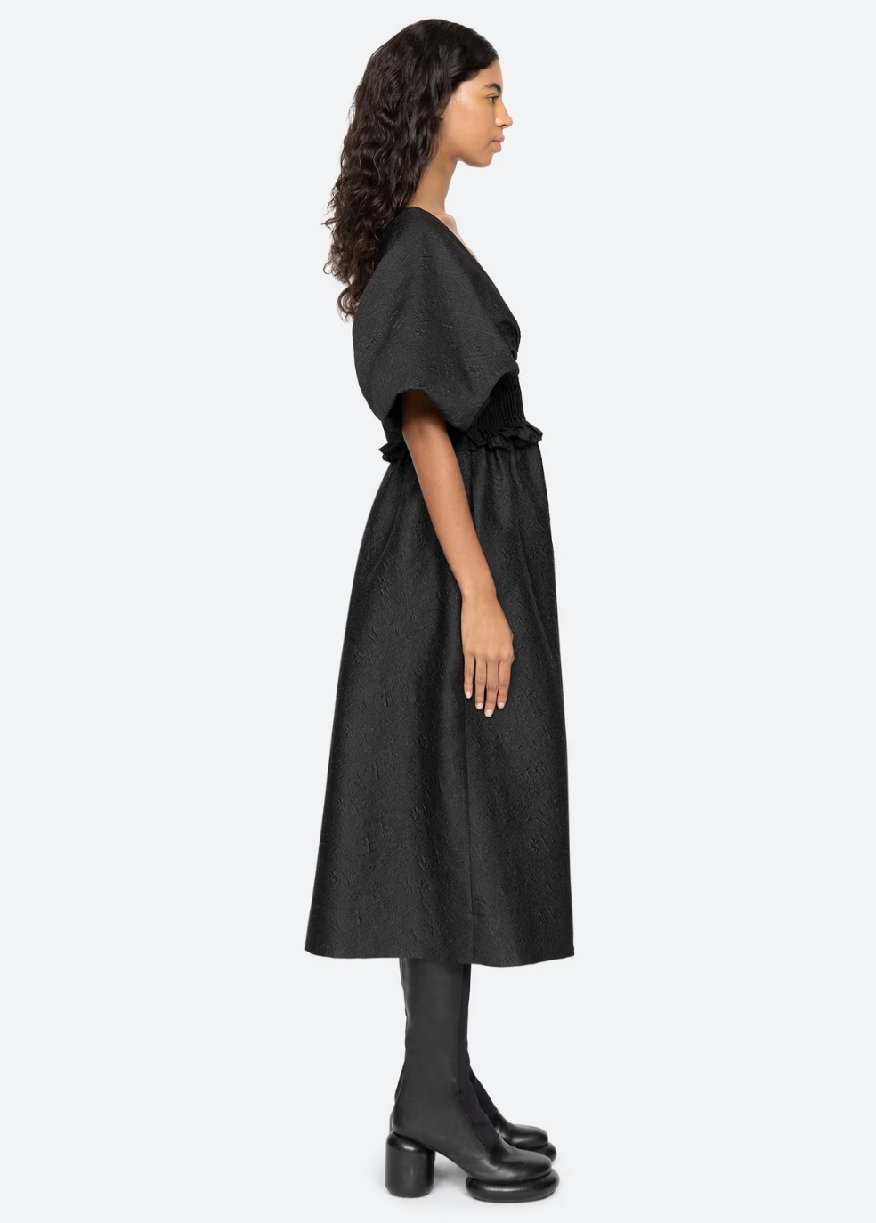 A woman with long curly hair stands in profile against a plain background, wearing the Elegi Solid Short Sleeve Dress by Sea New York. The black jacquard dress, featuring oversized puff sleeves, falls to her knees. She also pairs it with black mid-calf boots with a block heel.