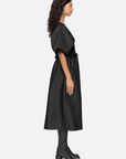 A woman with long curly hair stands in profile against a plain background, wearing the Elegi Solid Short Sleeve Dress by Sea New York. The black jacquard dress, featuring oversized puff sleeves, falls to her knees. She also pairs it with black mid-calf boots with a block heel.