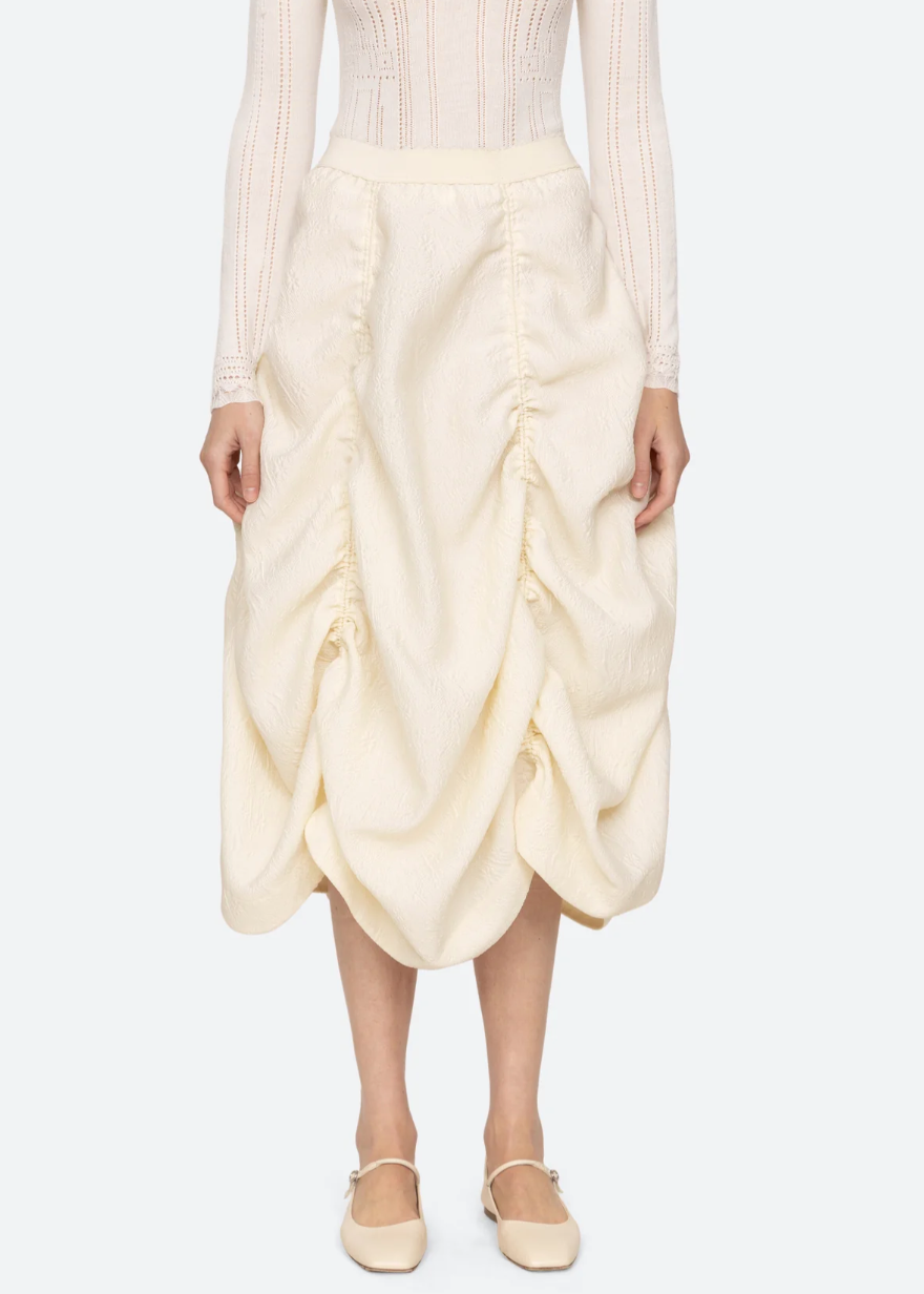 A person stands facing forward wearing a Sea New York Elegi Solid Skirt with ruching details down the sides, paired with a long-sleeved, knit top in a matching color. The outfit is complemented by cream-colored, closed-toe, flat shoes.