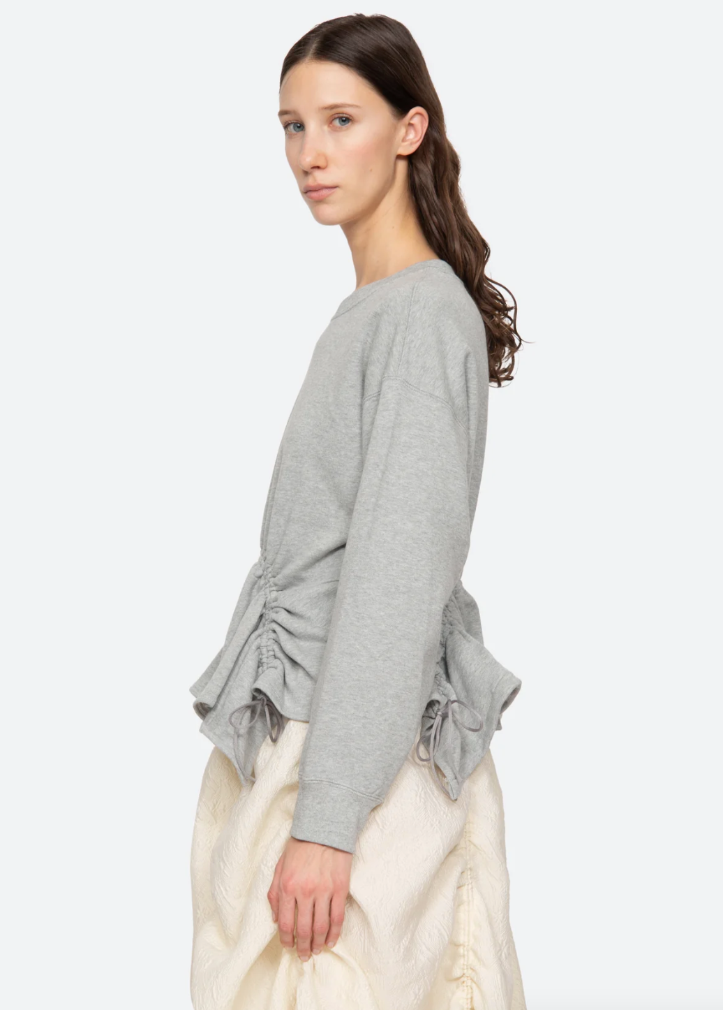 A person with long brown hair stands in side profile, showcasing the Sea New York Elegi Solid Long Sleeve Sweatshirt, featuring gathered sides and an asymmetrical hem. They pair it with light-colored textured pants for a sophisticated look. The relaxed fit and neutral expression against a plain white background complete the understated ensemble.