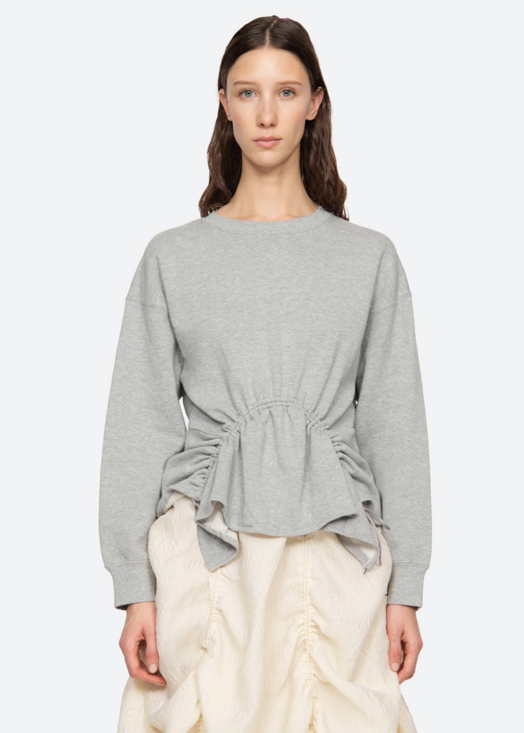 A woman with long brown hair stands against a plain background, wearing the Elegi Solid Long Sleeve Sweatshirt by Sea New York, which features adjustable drawstring cords at the waist. She pairs it with a cream-colored skirt. The relaxed fit sweatshirt has long sleeves, and she is looking directly at the camera with a neutral expression.