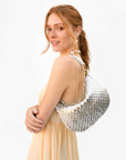 A person in a light beige dress is smiling, looking over their shoulder. They are carrying the Clare Vivier Petite Moyen S24, a woven metallic handbag with crossbody straps, and wearing pearl drop earrings, standing against a plain white background.