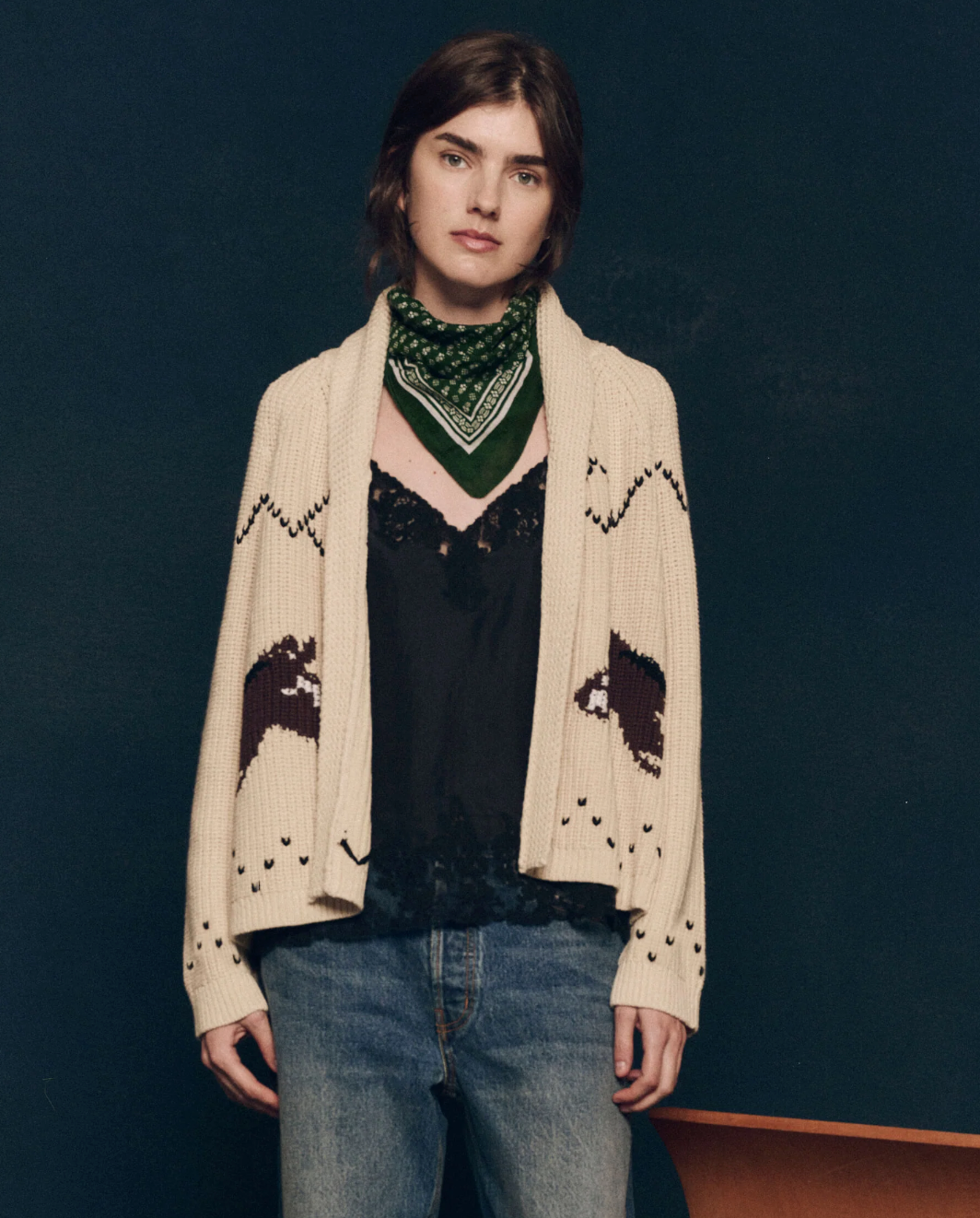 A person with short brown hair wearing The Great Inc.'s The Horse Lodge Cardigan, adorned with hand-embroidered horses, a green bandana around their neck, a black lace top, and blue jeans stands against a dark background.