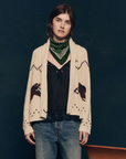 A person with short brown hair wearing The Great Inc.'s The Horse Lodge Cardigan, adorned with hand-embroidered horses, a green bandana around their neck, a black lace top, and blue jeans stands against a dark background.