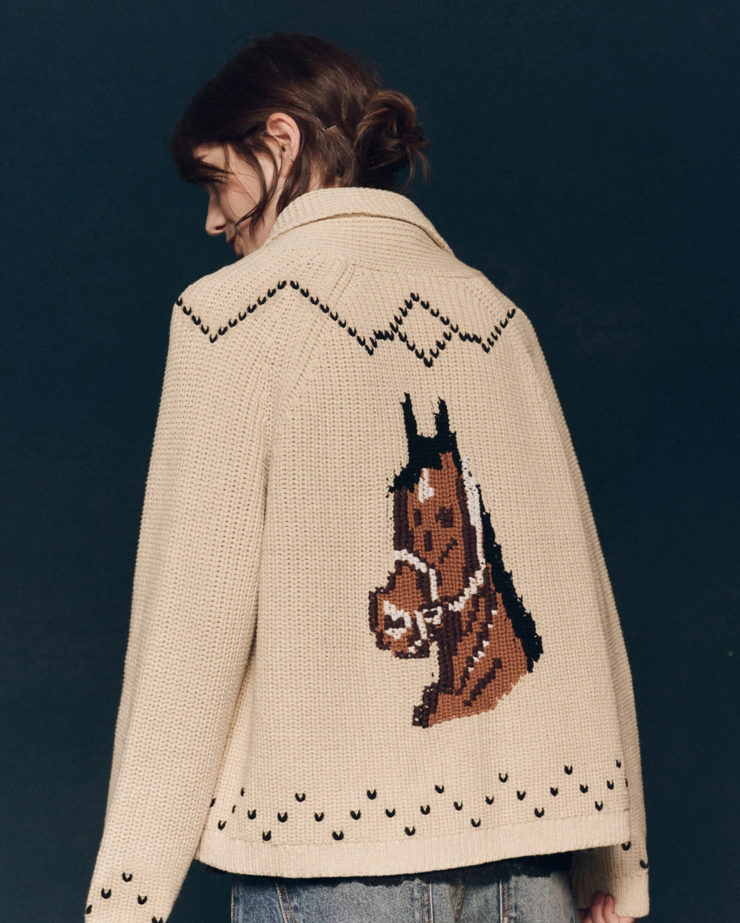 A person with dark hair is turned away, wearing The Great Inc.'s Horse Lodge Cardigan made from a cotton blend yarn, featuring a decorative pattern and an impressive large image of hand-embroidered horses on the back. The horse design includes detailed stitching, adding a rustic, country-inspired touch to the garment.