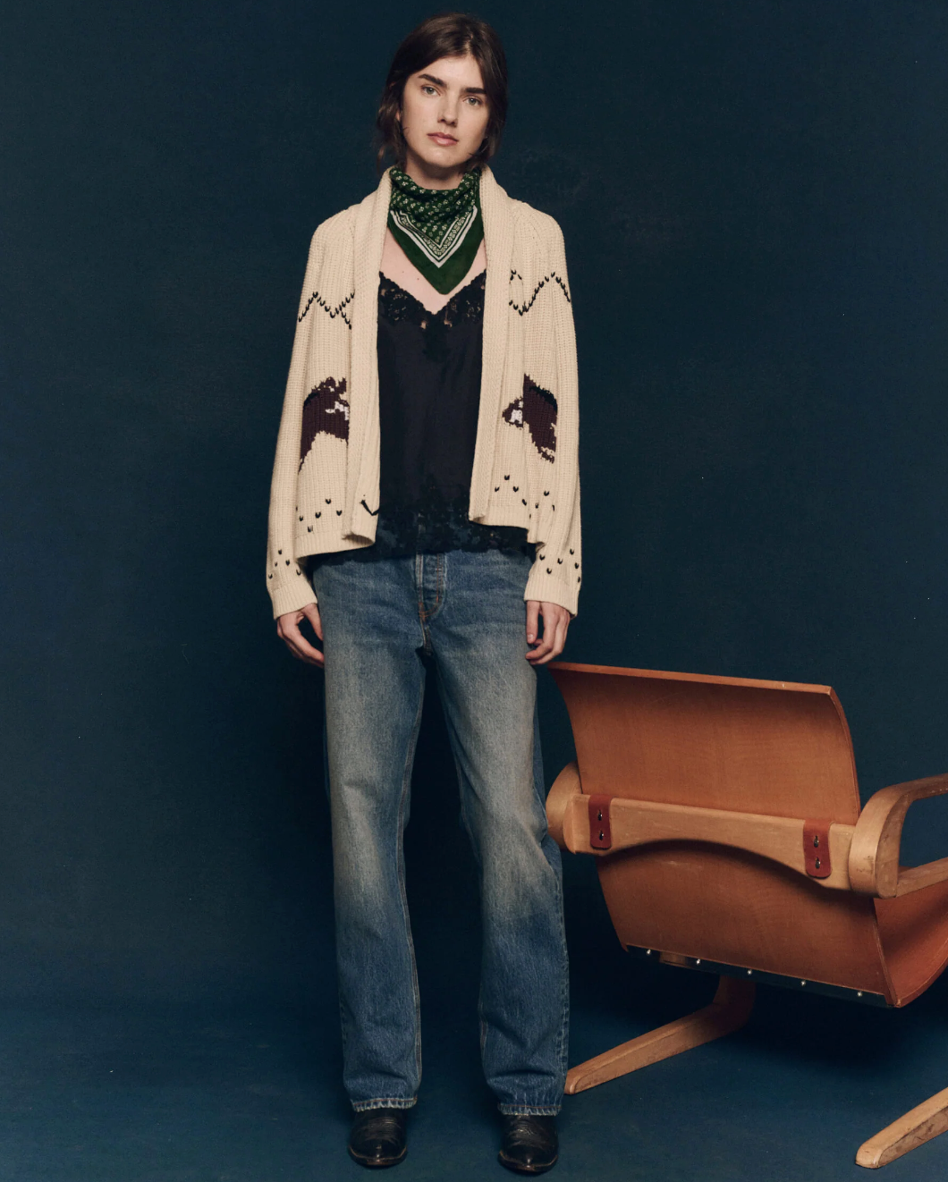 A person with shoulder-length hair stands in front of a dark background, wearing The Great Inc.'s The Horse Lodge Cardigan, which features hand-embroidered horses. They also sport a green bandana, a black top, and blue jeans. To their right is a wooden chair that adds a touch of western nostalgia to the scene.