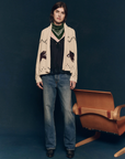 A person with shoulder-length hair stands in front of a dark background, wearing The Great Inc.'s The Horse Lodge Cardigan, which features hand-embroidered horses. They also sport a green bandana, a black top, and blue jeans. To their right is a wooden chair that adds a touch of western nostalgia to the scene.