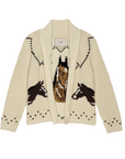 The Horse Lodge Cardigan by The Great Inc. is a cream-colored, knitted piece with a shawl collar. It features an exquisite design of three hand-embroidered brown and white horses on the front and has a textured, cozy appearance accented with black detailing around the shoulders and hem.