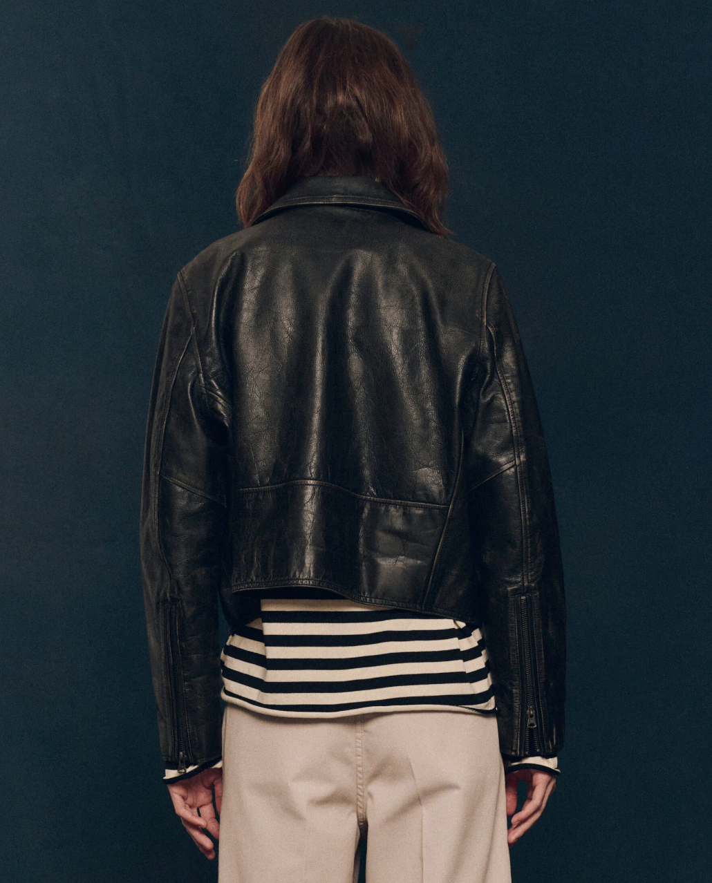 A person with long hair is photographed from behind, wearing The Perfect Leather Jacket by The Great Inc., over a black and white striped shirt and beige pants, standing against a dark background.