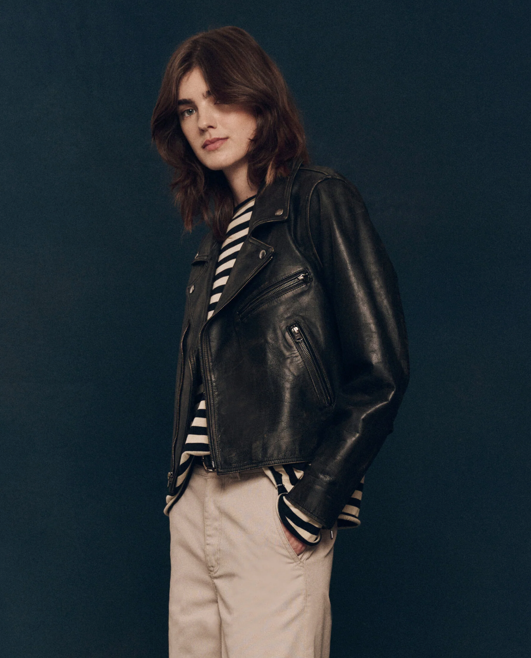 A person with shoulder-length brown hair stands against a dark background. They are wearing The Great Inc.'s Perfect Leather Jacket with antique silver snaps over a black and white striped shirt, paired with light beige pants. They are looking directly at the camera.