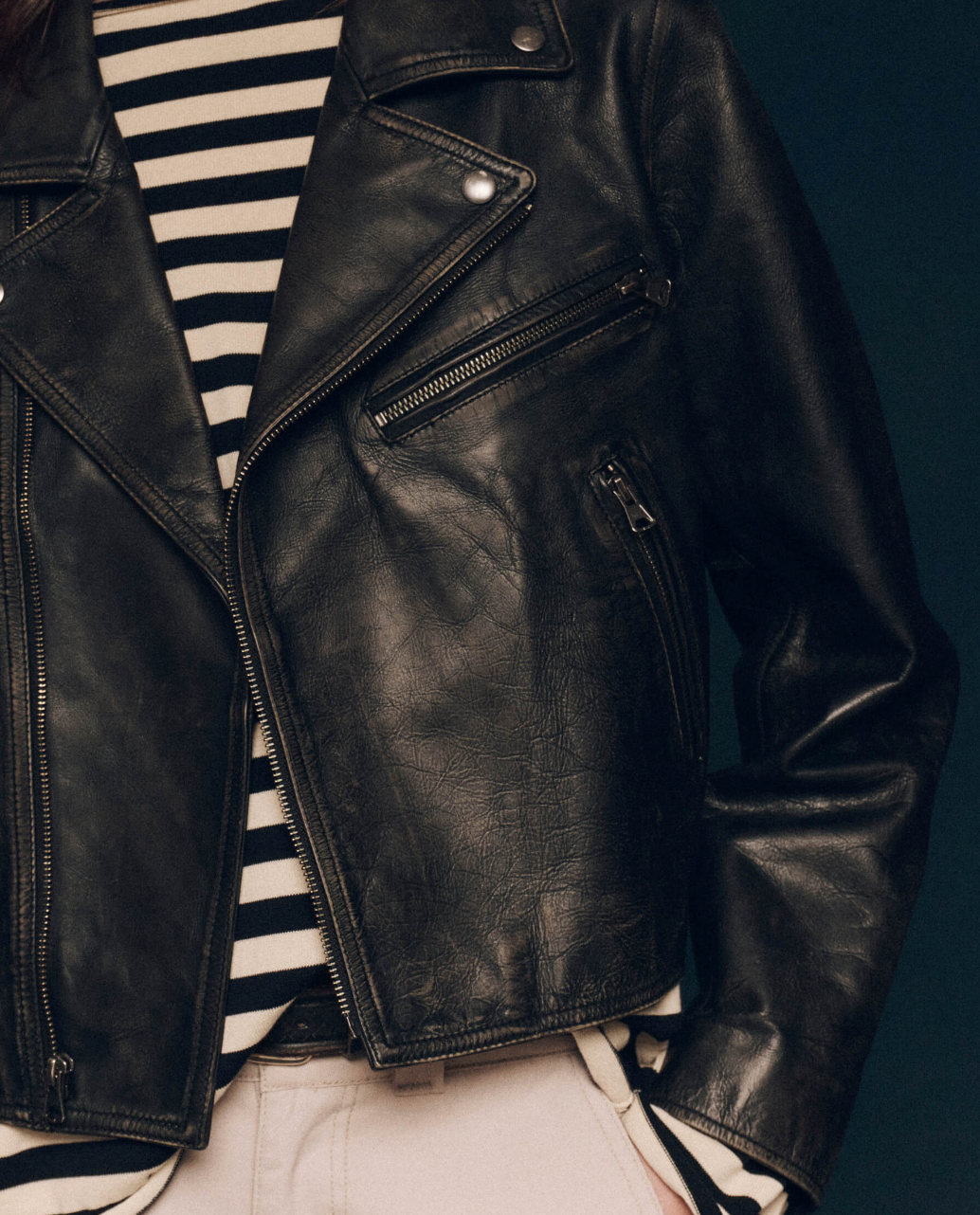 Close-up of a person wearing The Perfect Leather Jacket by The Great Inc. over a black and white striped shirt. The cow leather jacket features multiple zippers, antique silver snaps, and buttons. The bottom of the shirt is partially visible, and the person's hands are in the jacket pockets. The background is dark.