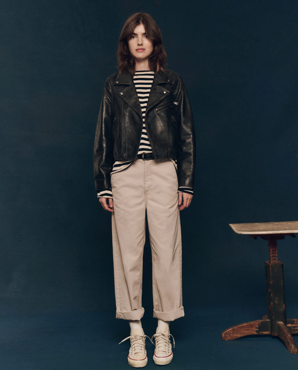 A person with shoulder-length brown hair stands against a dark background, sporting The Perfect Leather Jacket from The Great Inc., featuring antique silver snaps. They wear a black and white striped shirt, beige cuffed pants, and white sneakers. Beside them is a wooden stool. The person is looking directly at the camera.