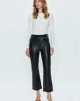 A person with long, wavy hair stands facing forward against a plain white background. They are wearing a white long-sleeved shirt, the buttery soft black leather Lennon Pant by Pistola, and black heeled shoes. Their arms are relaxed at their sides.