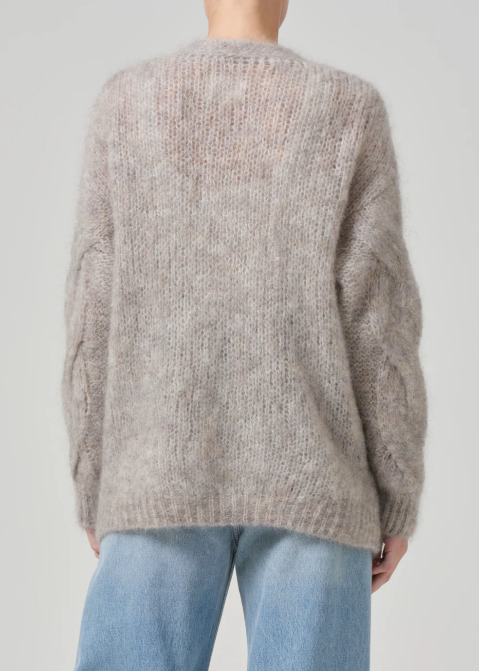 A person wearing a Citizens Of Humanity/AGOLDE padma cable cardigan, which boasts a soft, fuzzy beige mohair blend with loose, long sleeves and a chunky knit texture, paired with blue jeans is seen from the back. The cardigan has a relaxed fit and the background is plain light gray.