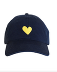 The Embroidered Heart Hat by Kerri Rosenthal, a quintessential collegiate-style navy blue baseball cap, showcases a small yellow heart embroidered at the front center. It features a curved brim and noticeable stitching along the panels and edges against a plain white background.