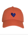 Introducing the Kerri Rosenthal Embroidered Heart Hat, featuring a blue heart embroidered on the front center. Part of the KR CAP-Sule Collection, this orange baseball cap is made from 100% cotton and includes a curved brim with visible stitching lines, set against a plain white background.