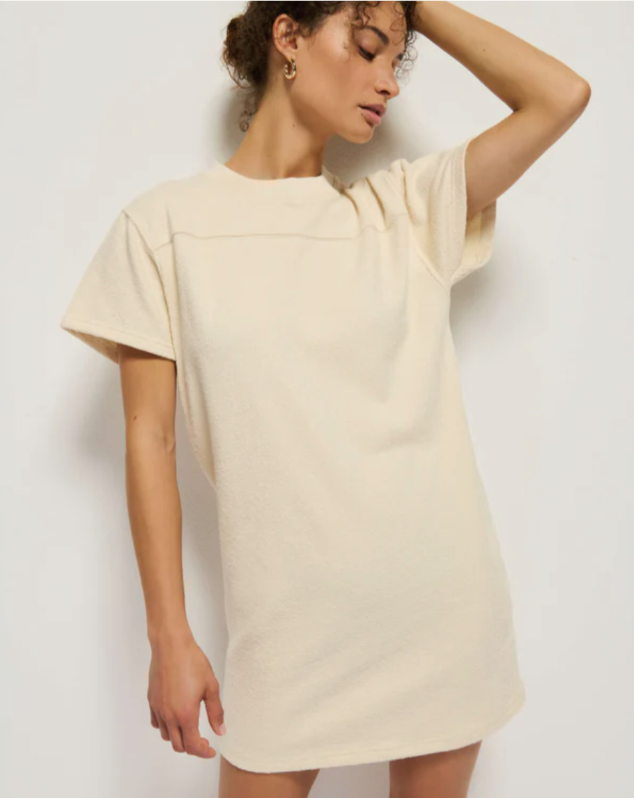 A person with curly hair stands against a plain white background, wearing the Finn Crewneck Dress by Nation LTD. The beige T-shirt dress features short sleeves and a crew neck. The individual has one arm raised, resting their hand on their forehead, and is looking down to the side. This stylish dress is also machine washable for easy care.