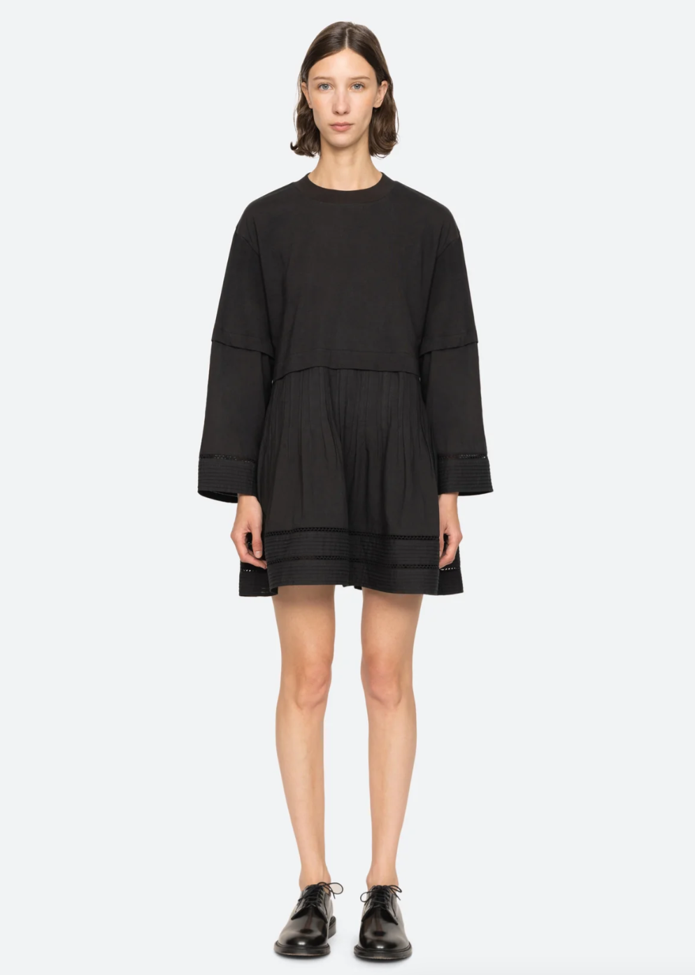 A person with short, dark hair is wearing a black Renata Tunic Dress by Sea New York, featuring a relaxed fit and pleated detailing. They are standing against a plain white background and are also wearing black oxford shoes.