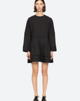 A person with short, dark hair is wearing a black Renata Tunic Dress by Sea New York, featuring a relaxed fit and pleated detailing. They are standing against a plain white background and are also wearing black oxford shoes.