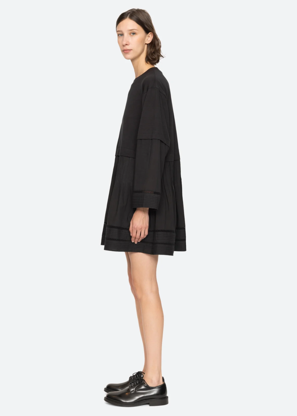 A person with short, dark hair stands in profile, wearing a black Renata Tunic Dress by Sea New York, featuring pleated detailing at the hem. The relaxed fit complements their shiny black oxford shoes as they stand against a plain, light gray background.