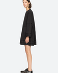 A person with short, dark hair stands in profile, wearing a black Renata Tunic Dress by Sea New York, featuring pleated detailing at the hem. The relaxed fit complements their shiny black oxford shoes as they stand against a plain, light gray background.