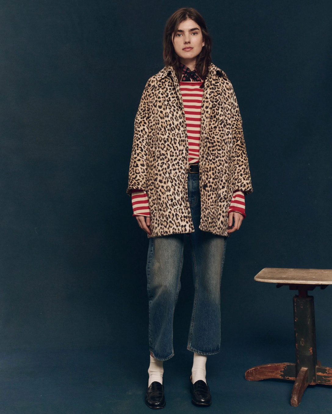 A person with shoulder-length hair stands against a dark background wearing The Car Coat, a midcentury-inspired faux fur leopard print piece from The Great Inc., over a red and white striped shirt, blue cropped jeans, white socks, and black loafers. There is a small wooden stool to the right.