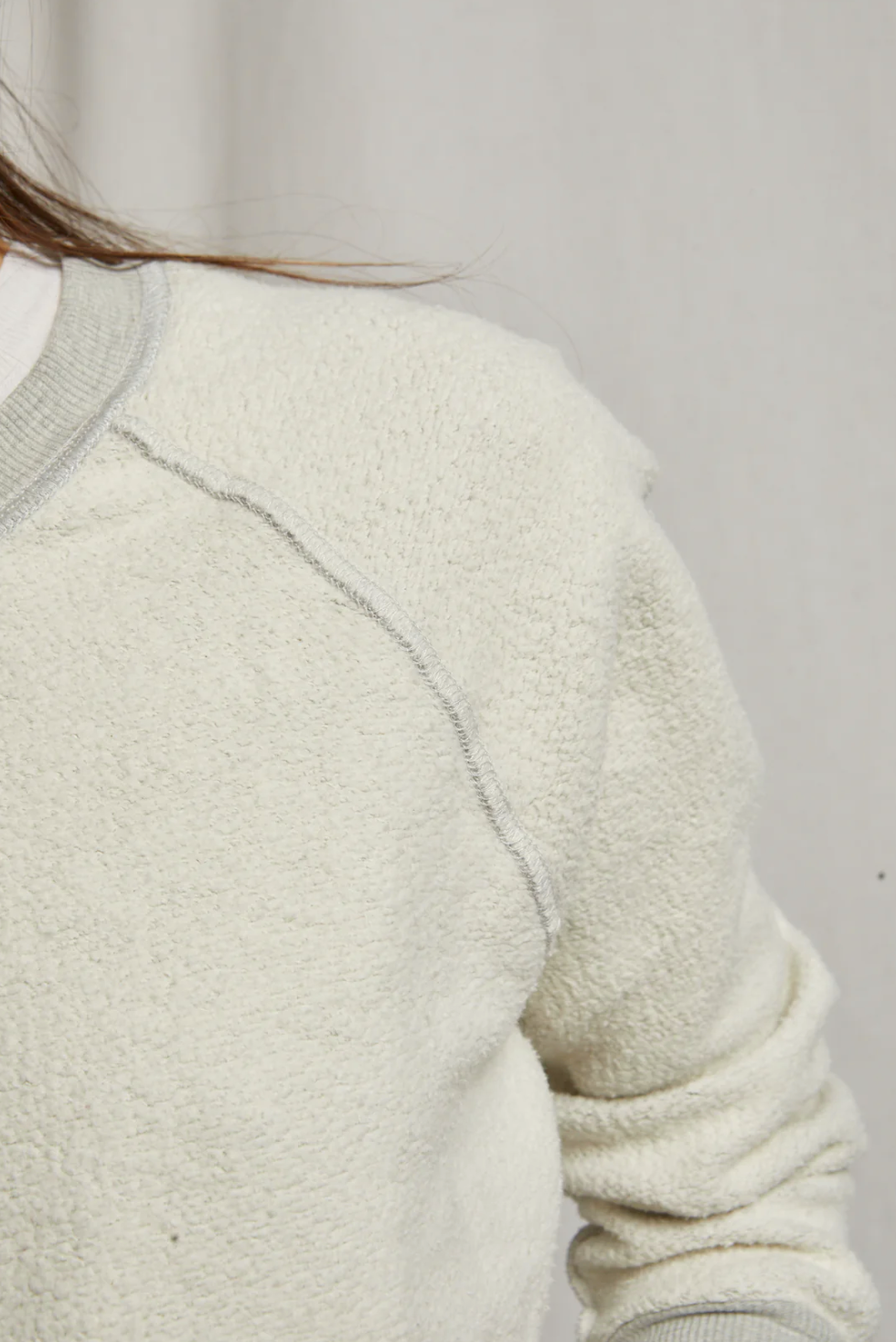 Close-up of a person wearing the Ziggy Inside Out LS Crewneck Sweatshirt by Perfectwhitetee, exuding cozy essential vibes with its cream-colored textured fabric. Light gray stitching is visible along the shoulder and sleeve seams. The partially visible upper body highlights the buttery soft fleece on the shoulders and sleeves against a plain light gray background.