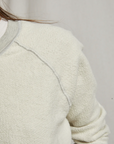 Close-up of a person wearing the Ziggy Inside Out LS Crewneck Sweatshirt by Perfectwhitetee, exuding cozy essential vibes with its cream-colored textured fabric. Light gray stitching is visible along the shoulder and sleeve seams. The partially visible upper body highlights the buttery soft fleece on the shoulders and sleeves against a plain light gray background.