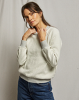 A person with long hair is wearing a cozy Ziggy Inside Out LS Crewneck Sweatshirt by Perfectwhitetee, posing with hands near the neckline, and blue jeans, stands in front of a plain light-gray backdrop.