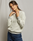 A person with long dark hair stands against a light backdrop, wearing the Ziggy Inside Out LS Crewneck Sweatshirt by Perfectwhitetee paired with blue jeans. They have a contemplative expression, and their hands are gently touching their collarbone and neck.