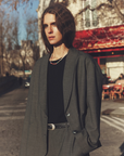 A person with long hair is wearing the Jonah Jacket from Xirena, a grey wool blazer with welt pockets and matching pants. They are standing outdoors on a sunny day, with a red awning and outdoor seating from a café in the background, along with trees and buildings.