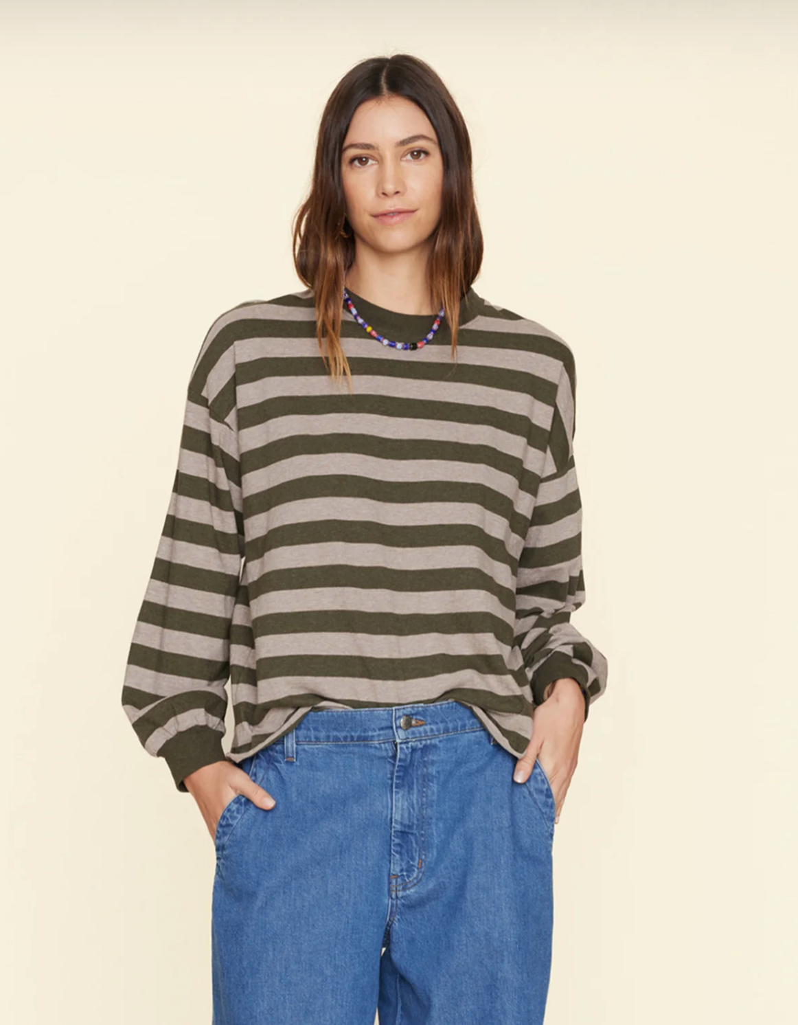 A person with long brown hair stands against a plain backdrop, wearing a green and grey wide-striped Honor Tee by Xirena and blue jeans. Their hands are in their pockets, and they have a relaxed expression.