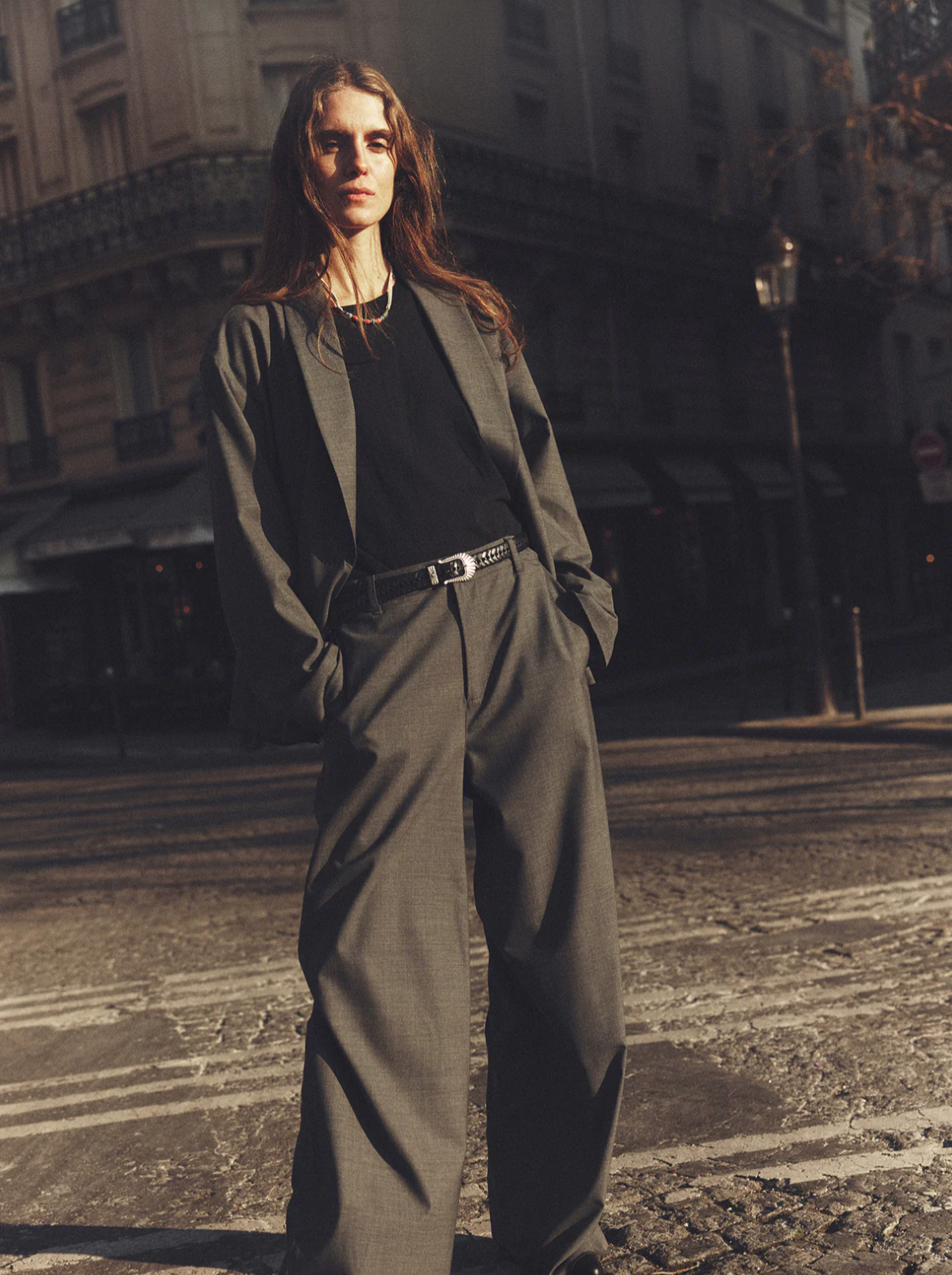 A person with long hair stands confidently on a city street, wearing the loose-fitting grey Portlyn Pant by Xirena paired with a black shirt. They have their hands in their pockets and the sun casts a warm light, creating dramatic shadows. The background shows a building and blurred street details.