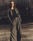 A person with long hair stands confidently on a city street, wearing the loose-fitting grey Portlyn Pant by Xirena paired with a black shirt. They have their hands in their pockets and the sun casts a warm light, creating dramatic shadows. The background shows a building and blurred street details.